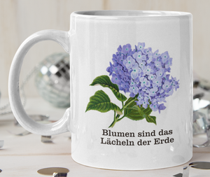 Tasse Weiß bedruckt mit Spruch: Blumen sind das Lächeln der Erde. Farbe der Blumen: Blau/Lila
